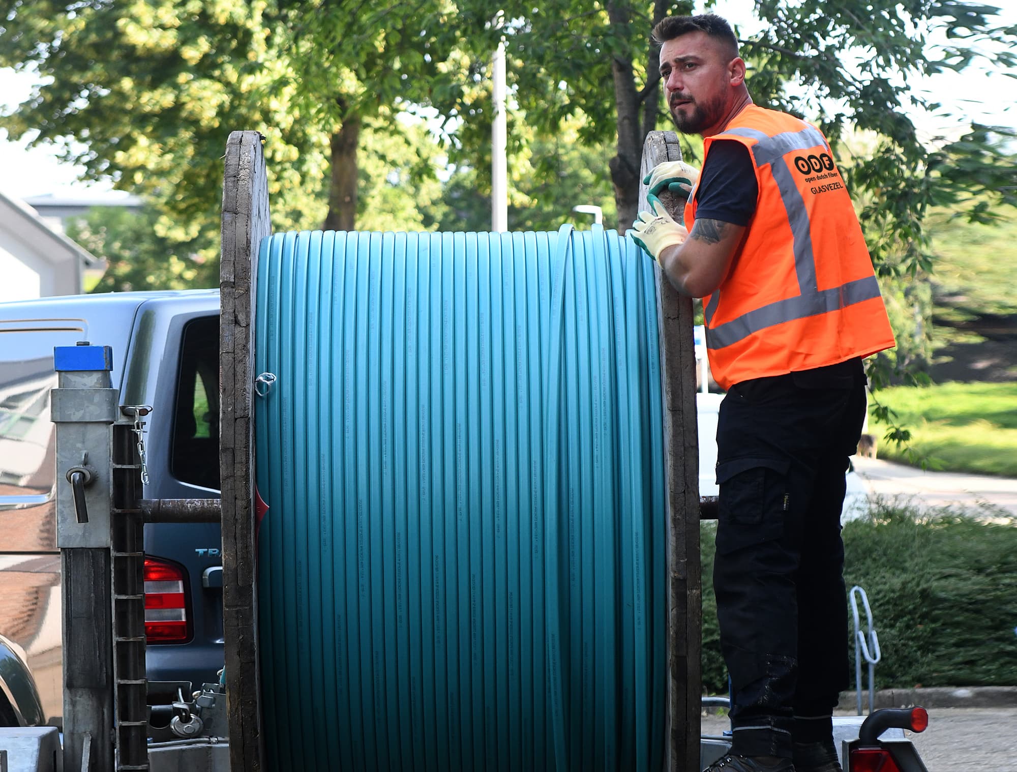 Werknemer in oranje veiligheidsvest die een grote spoel blauwe kabel verwerkt op een aanhangwagen naast een bestelwagen in een met bomen omzoomde wijk.